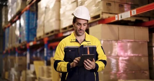 Warehouse worker in hardhat and safety jacket checks inventory data using tablet while walking
