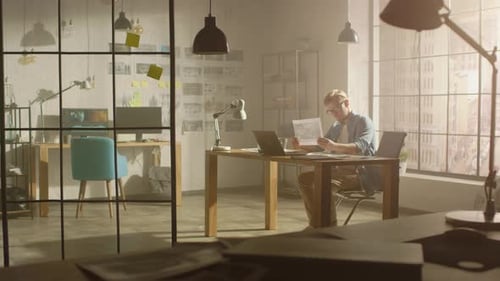 Professional Creative Designer Works on a Laptop in Loft Office, Looks at the Product Sketches and