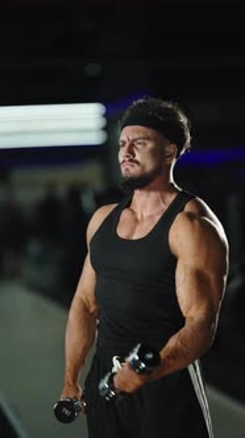 Focused Athlete Performing Intense Dumbbell Bicep Curls in a Dimly Lit Gym Showcasing Muscular Arms