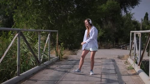 Joyful Woman Walking Dancing Along Listening Music in Wireless Headphones Outdoors on the Bridge