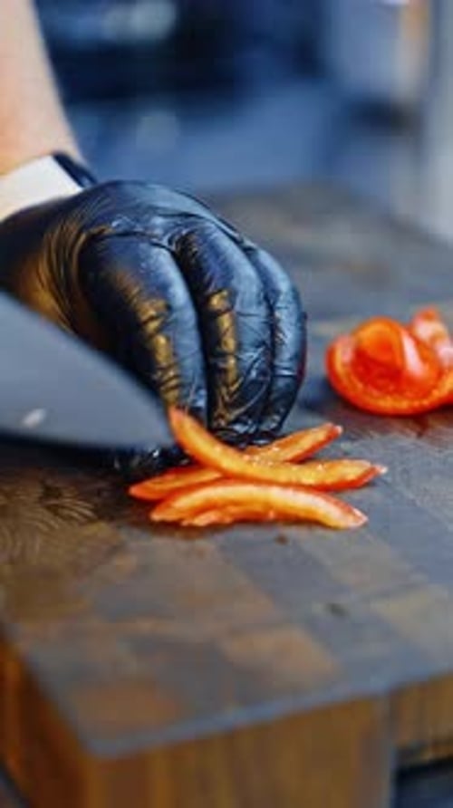 Male cook wearing black gloves cutting red pepper with a big knife. Salad preparation close up.
