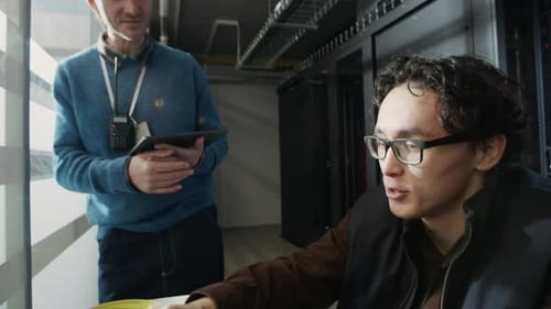 Two Network Engineers Monitoring Systems Using Laptop in Data Center Corridor