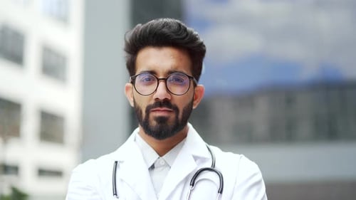 Close up portrait of a young confident doctor in a white coat and glasses standing outside in front