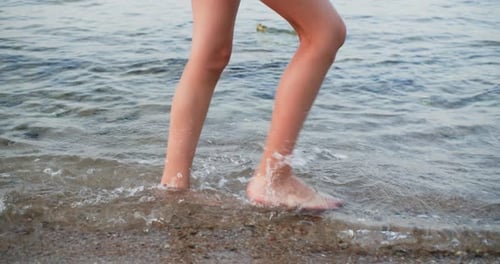 Slim Woman's Legs Walking Along Sea Water with Small Waves on Rocky Beach