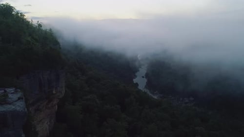 Luftbild, das zur blauen Stunde an einer Klippe zum nebligen Waldflusstal vorbeifliegt, 4K