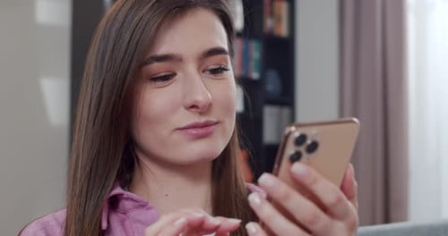 Close Up of Happy Caucasian Young Woman Sitting on Sofa and Chatting By Messages on Smartphone At