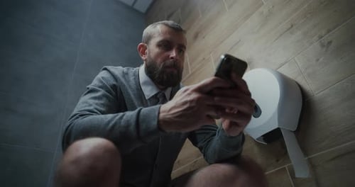 Businessman Using Smartphone Sitting on the Toilet in Restroom