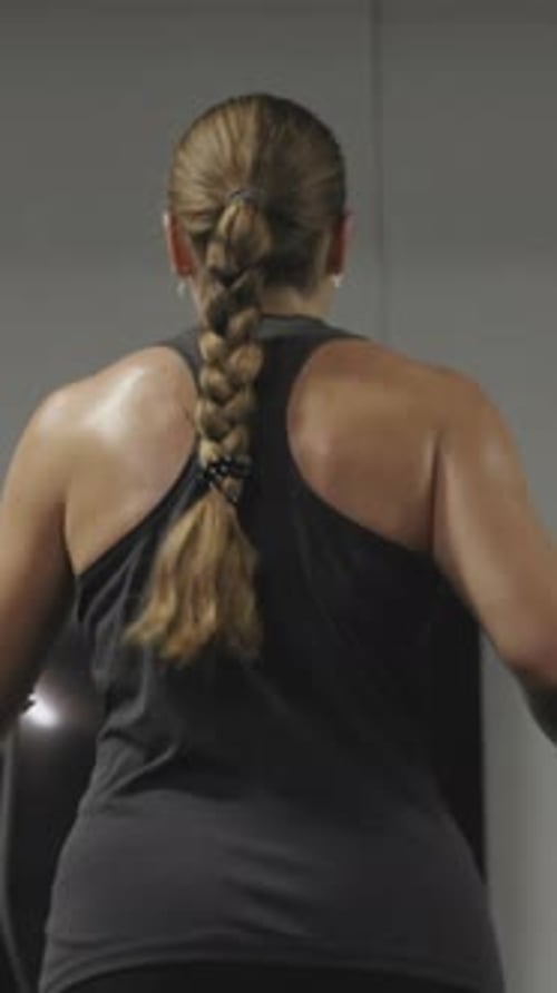 Vertical video. Sweat trickles down the back of a young woman working out on Elliptical Trainer.