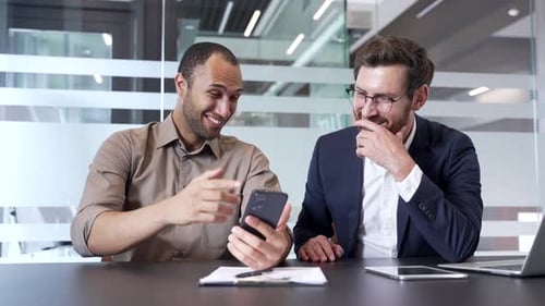 Two coworkers laughing looking at phone while sitting at desk at workplace in modern business office