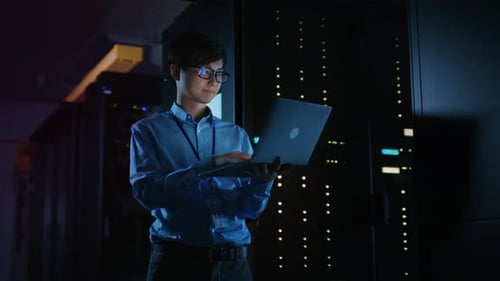 In Dark Data Center: Male IT Specialist Stands Beside the Row of Operational Server Racks, Uses Lap