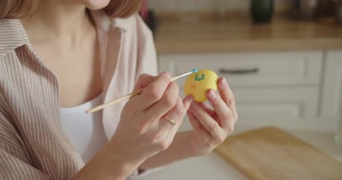 Young Woman Painting Easter Egg Sitting at the Table in Bright Modern Kitchen