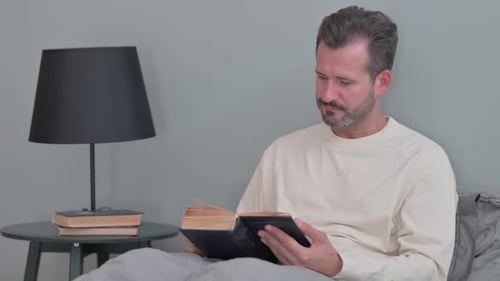 Man reads book in bed in bedroom