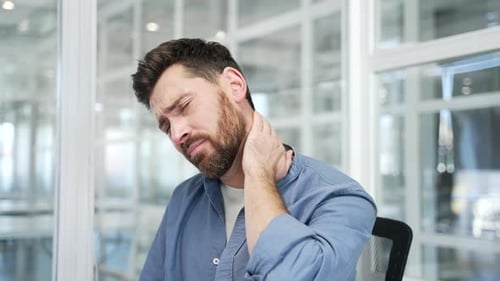 Tired businessman suffering from neck pain while sitting at desk at workplace in business office.