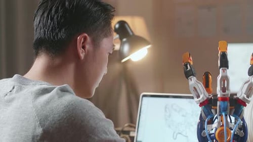 Over The Shoulder View Of A Male Designing A Cyborg Hand On A Laptop At Home