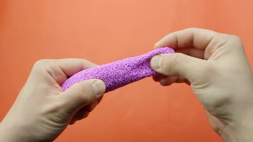 Hands tearing apart lilac floam slime with foam beads for ASMR play on vivid orange background