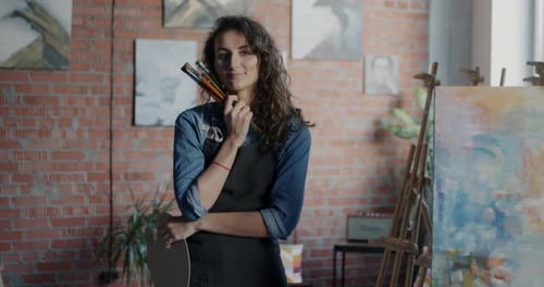 Portrait of Young Woman Artist in Apron Standing in Creative Studio Holding Paint Brushes Smiling