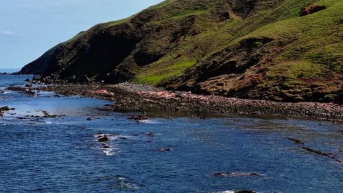 Wellen bewegen sich neben grünen Hügeln in Richtung steiniges Ufer, Meer rollt entlang eines zerklüfteten Abhangs auf felsigen Strand