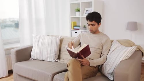 Leisure, Literature And People Concept - Indian Man Sitting On Sofa And Reading Book At Home