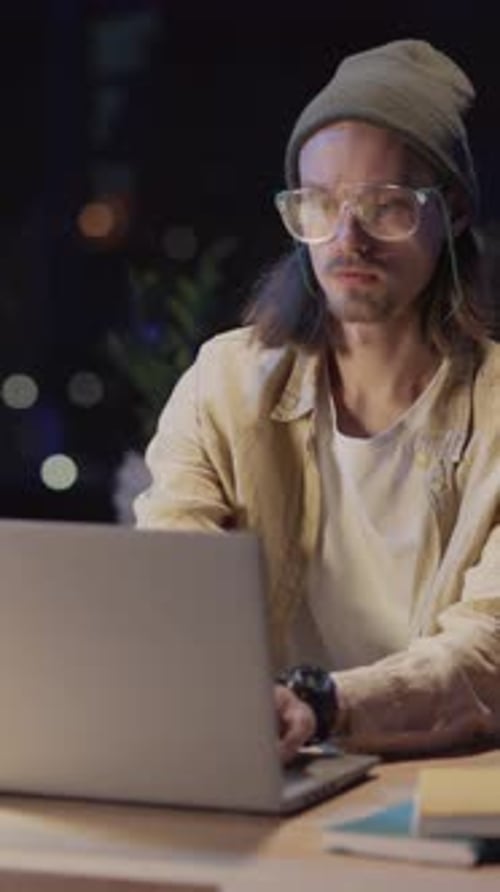 A Focused Man in Glasses and a Beanie Types on His Laptop Immersed