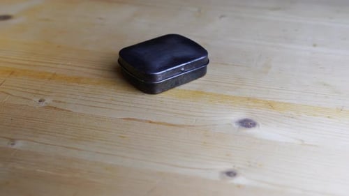Small silver metal box lies on a wooden table and rotates with dark me behind