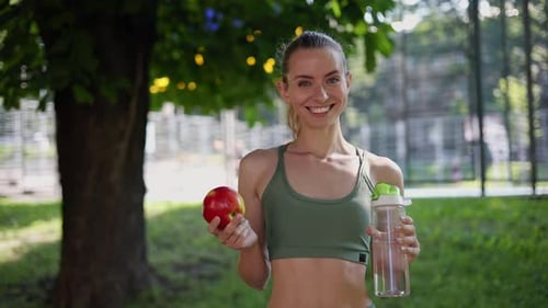 Smiling Woman Holding Apple and Water in Park