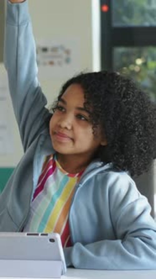 Vertical video: Noticing teacher prompt, girl in blue hoodie raising right hand in class, tablet