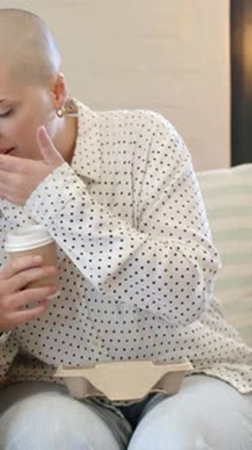 Vertical video: Sipping cup, woman in dotted blouse wiping mouth, reaching tray for cupcake on sofa