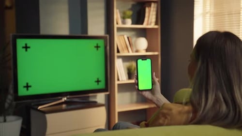 Woman on Sofa in Living Room Holding Smartphone with Blank Chroma Key Screen While Tv Also Shows