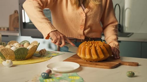 Woman Slicing Golden Bundt Cake On Wooden Board In The Kitchen, With Painted Easter Eggs Scattered