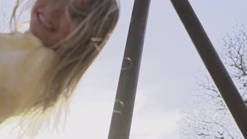 Happy Little Blonde Girl Looks at Smiling Camera While Swinging on Wooden Swing in Playground