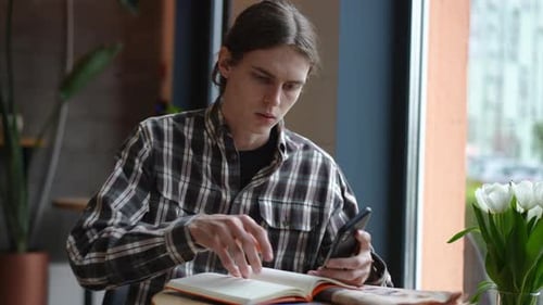 Concentrated Male Student Sitting in Cafe with Smartphone Writing with Pen in Slow Motion Portrait