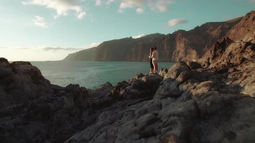 In a serene coastal setting, a couple embraces on rocky terrain overlooking the ocean, with steep cl