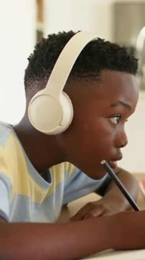 Vertical video: Leaning forward, kid in striped shirt doing homework with pencil at home in headset