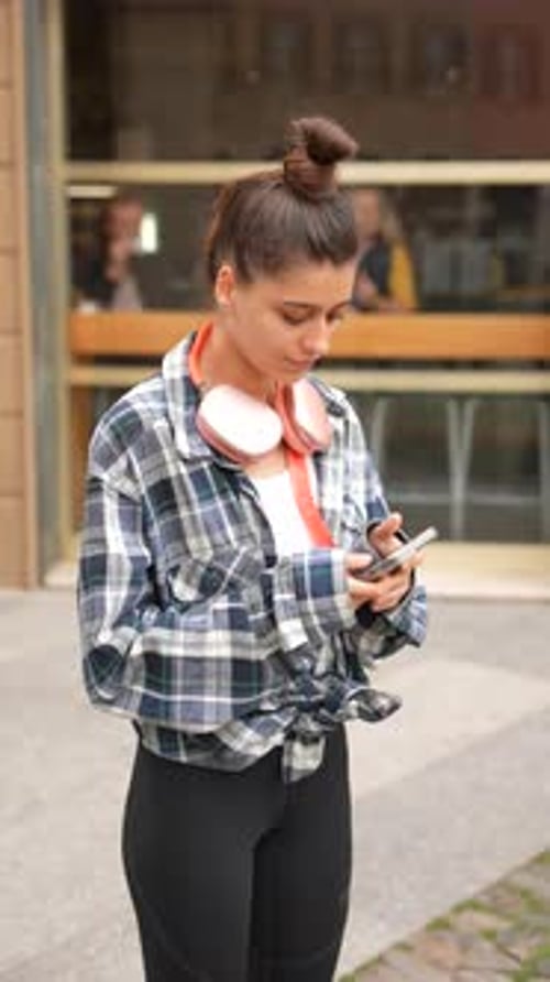 A Sporty Young Woman Wearing Headphones and Holding a Smartphone in the City Center