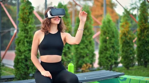 Smiling woman using VR headset in a tracksuit while sitting on a bench on a sports field in a park