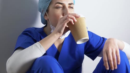 Tired Woman Doctor Drinks Coffee in Blue Scrubs
