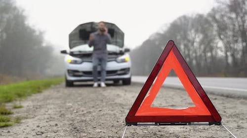 Road Side Warning Triangle Warning Oncoming Traffic of a Broken Down Car with a Man Using His Cell