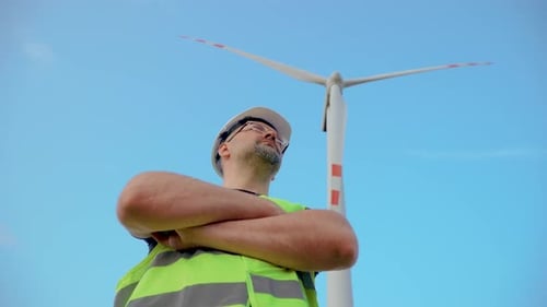 Portrait of Confident Man Engineer in Protective Helmet and Uniform in Front of Rotating Windmill