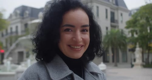 A Portrait of a Young Woman in a Cozy Coat Smiling and Happy Standing in the Park By a Fountain