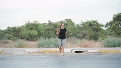 A young woman crosses the road, cars pass by, trees behind her