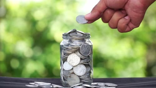 Human hand put and saving a coin in a glass container with full filled coins and blur green nature b
