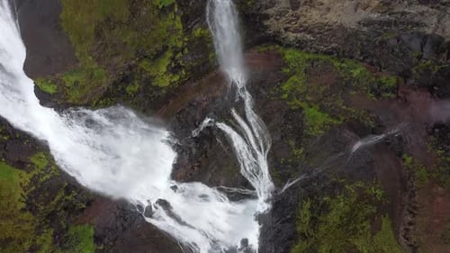 Aerial tilt up shot of powerful waterfalls between rocky and mossy mountains in Icelandic Scenery