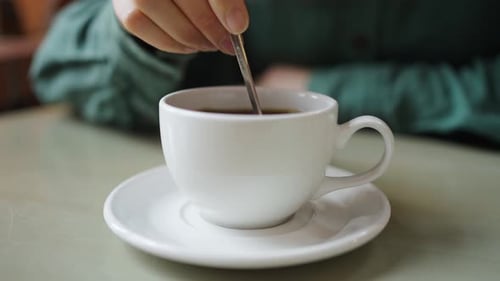 Closeup of Hands Gently Stirring a Steaming Cup of Coffee Placed on a Saucer on a Tabletop Capturing