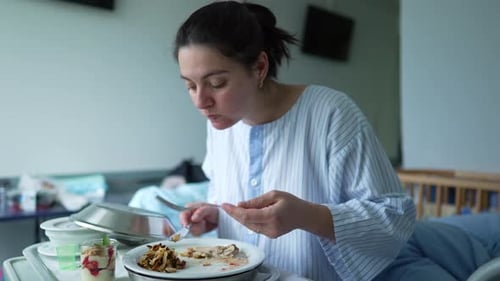 Woman in a hospital gown eating a meal on a tray while sitting on a hospital bed, showing the care
