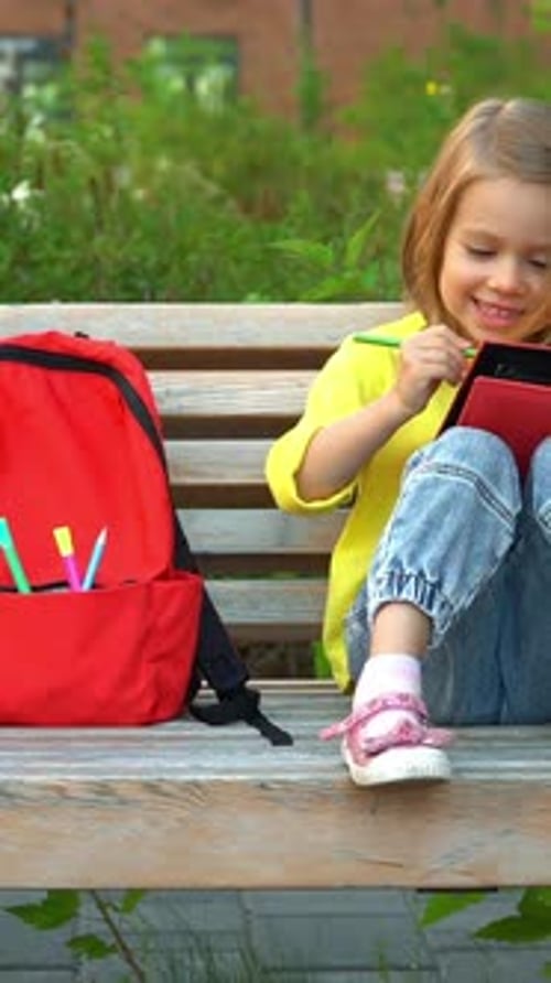 Little Schoolgirl Sits with Backpack in School Yard and Paint Outdoors Education