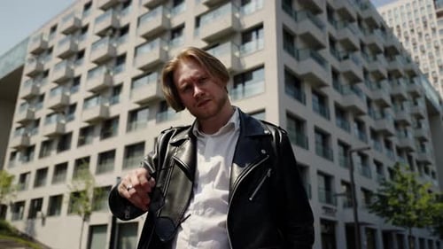 A Young Man Puts on Sunglasses and Looks at the Camera with a Serious Look Stylish Leather Jacket