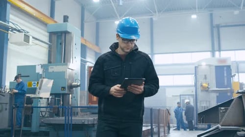 Um operário de fábrica com um capacete está andando e usando um tablet.