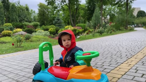 Lovely cute baby boy in jacket and hood stands near his toy car.