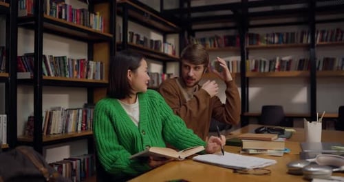 Happy Guy Talking to Girl While Studying in Front of Books at Table in Library