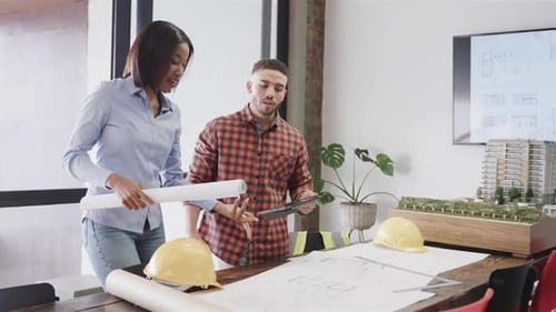 Diverse male and female architects with tablet discussing blueprints standing in office, slow motion